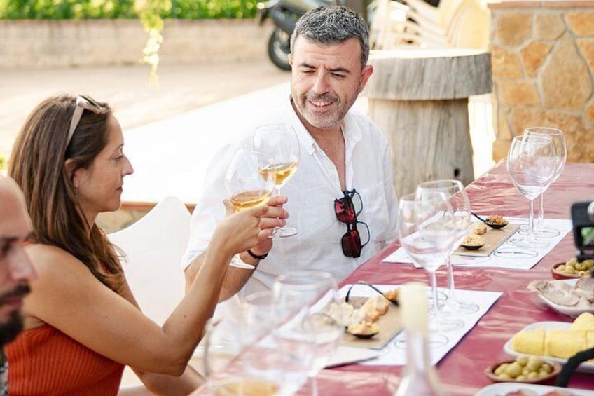 Un grupo de personas cenando al aire libre en una mesa, con copas de vino, platos de comida y un muro de piedra al fondo. 3