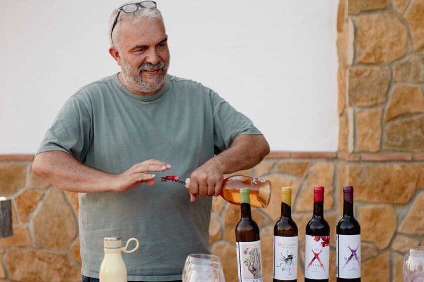 A man pouring a drink into a bottle with wine glasses and bottles in the foreground. 2