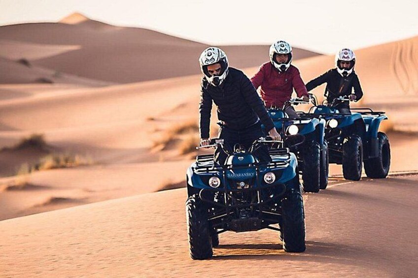 Drei Personen fahren mit Quads über hügeliges Sandgelände, im Hintergrund Dünen. 2