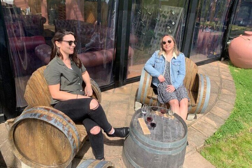 Two people sitting on wooden barrel chairs outdoors with a glass table between them. 5
