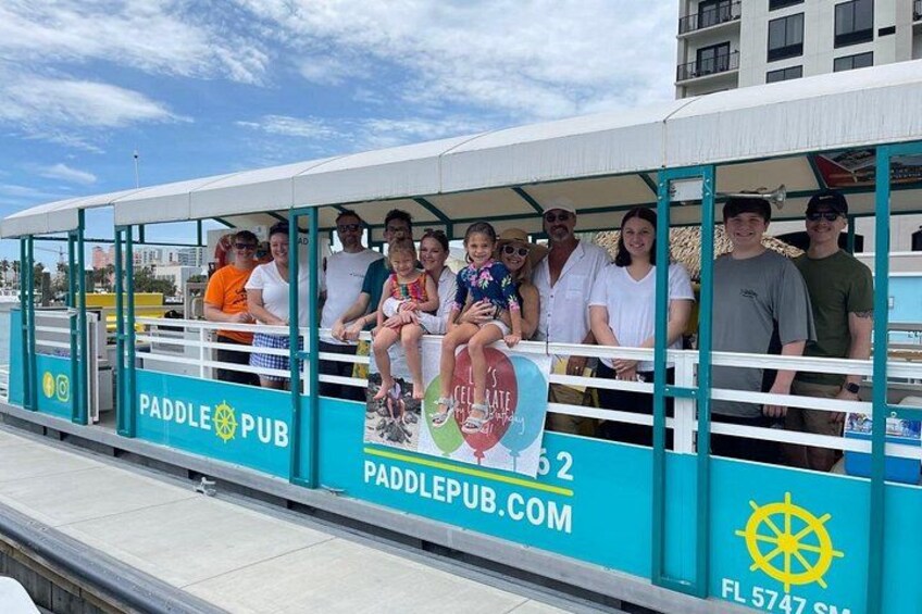 A group of people on a paddle boat with a sign that reads 'PADDLE PUB' and a website address. 1