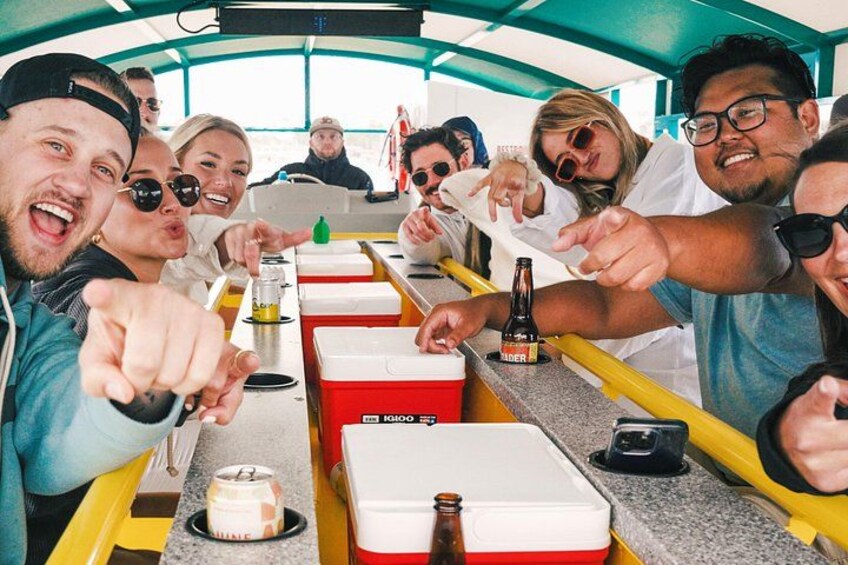 A group of people on a boat, some pointing at the camera, with a cooler, a bottle, and a jar on the table. 3
