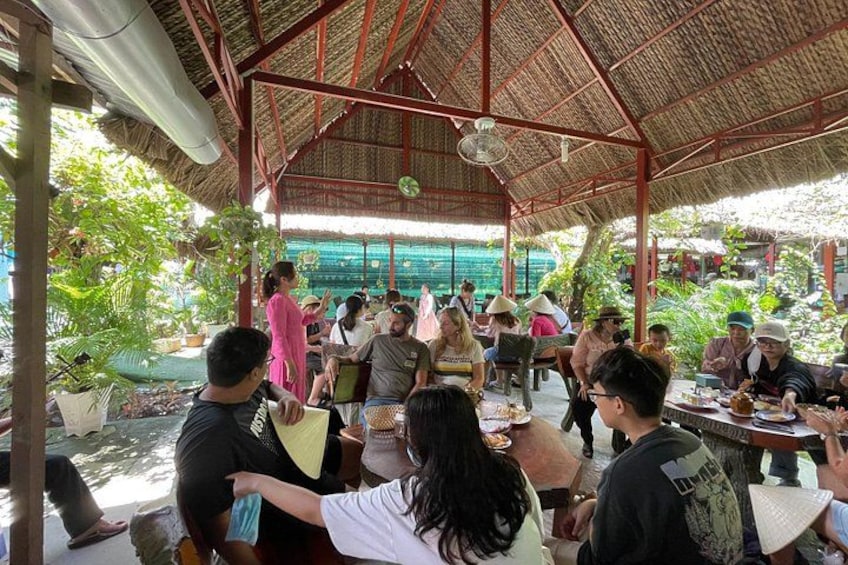 A group of people seated at tables under a thatched roof, engaged in conversation. 2