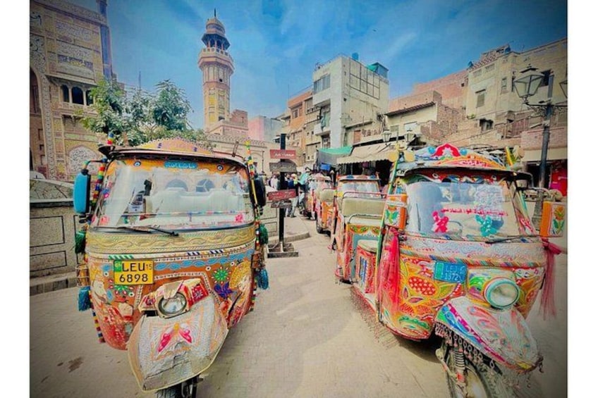 Colorful rickshaws parked on a street with buildings and a minaret in the background. 1
