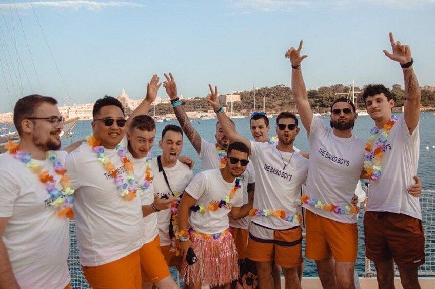 En gruppe mennesker på en båd, iført hvide t-shirts med logoet 'The Baja Boys' og orange shorts. 2