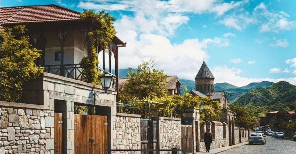 A stone-walled street with traditional buildings, a church with a tower, and mountains in the background. 3