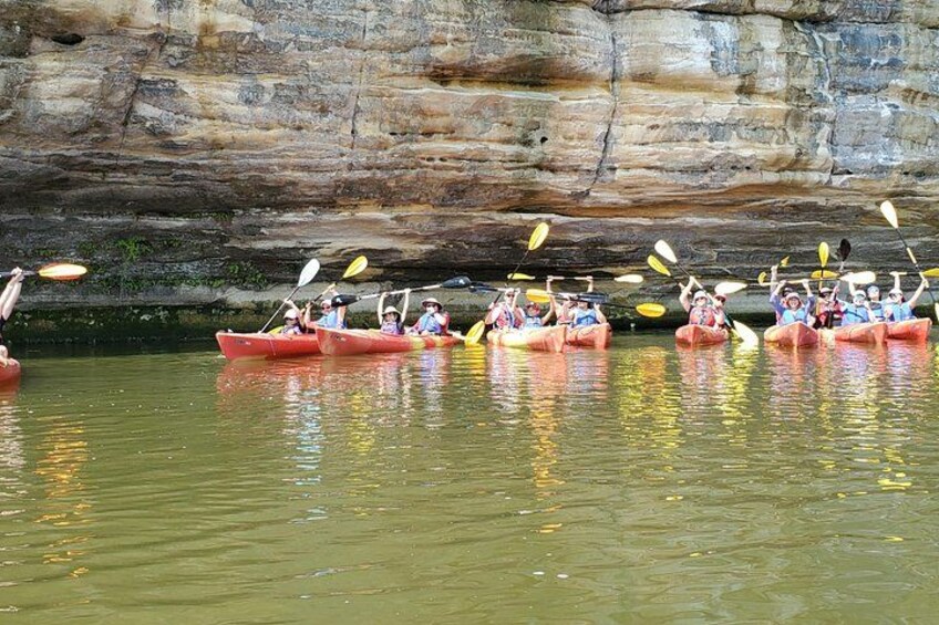 Eine Gruppe Menschen paddelt in roten Kanus auf einem ruhigen Fluss, im Hintergrund sind felsige Klippen zu sehen. 4