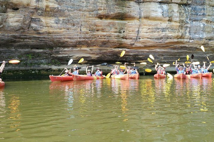 A group of people kayaking in red kayaks on a calm river with rocky cliffs in the background. 2