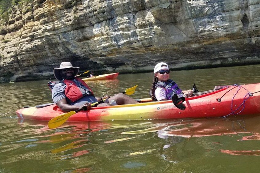 Two people kayaking on a calm river with rocky cliffs in the background. 3
