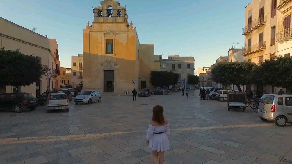 A woman walking towards a church with a bell tower, surrounded by parked cars and buildings. 4