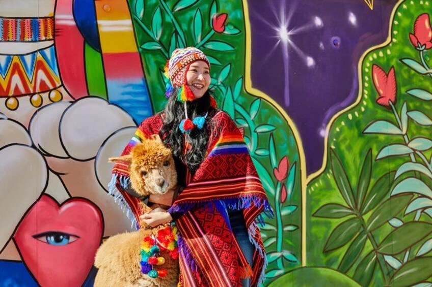 Een persoon in traditionele kledij met een lama, met een kleurrijke muurkunst op de achtergrond. 3