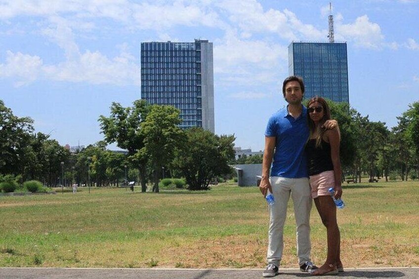Dos personas de pie en un parque con césped y árboles, y dos edificios altos al fondo. 5
