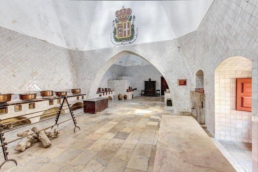 Ein Innenraum mit weiß gefliesten Wänden, einem Fliesenboden und einem Wappen an der Decke. Mehrere große Metallbehälter und ein Holztisch sind vorhanden. 4