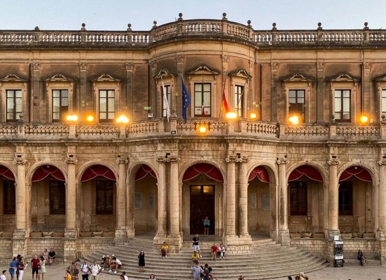 A historic building with classical architecture, featuring a central entrance with steps, arched windows, and flags. 3