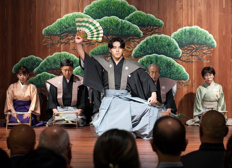 A traditional Japanese performance with actors in period costumes on a stage with a backdrop of stylized trees. 1