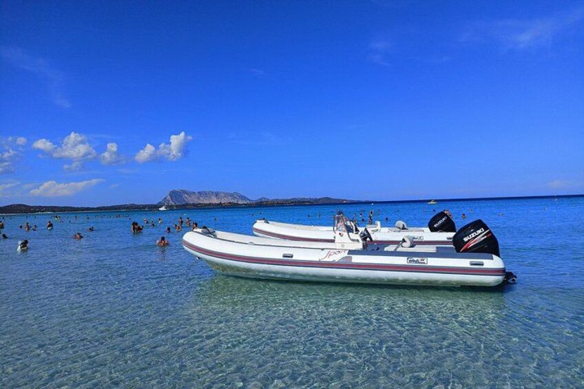 A clear blue sea with a boat docked near the shore, people swimming in the water, and a mountain in the background. 5