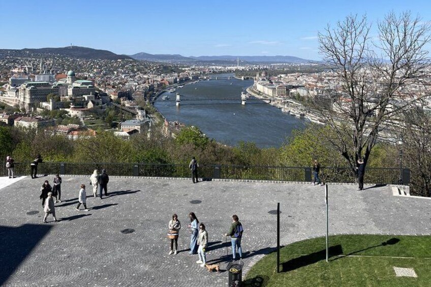 A riverside view with a bridge, a historic building complex, and people walking. 1