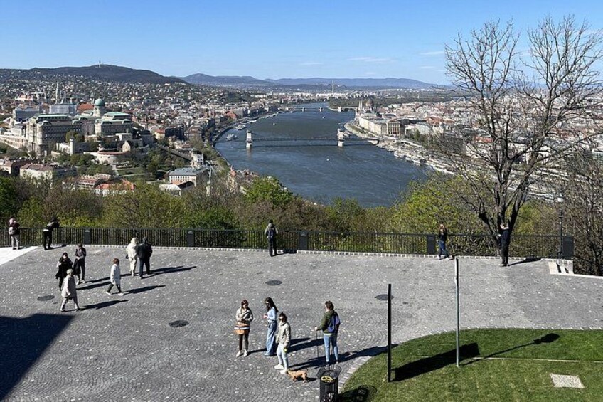 Blick auf einen Fluss mit einer Brücke, einem historischen Gebäude und Spaziergängern. 2