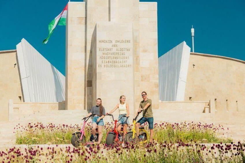 Un monumento con inscripciones, rodeado de un campo de flores, y una bandera al fondo. 4