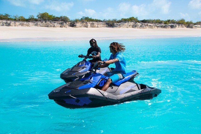 Two people riding jet skis on turquoise water with a beach and vegetation in the background. 2