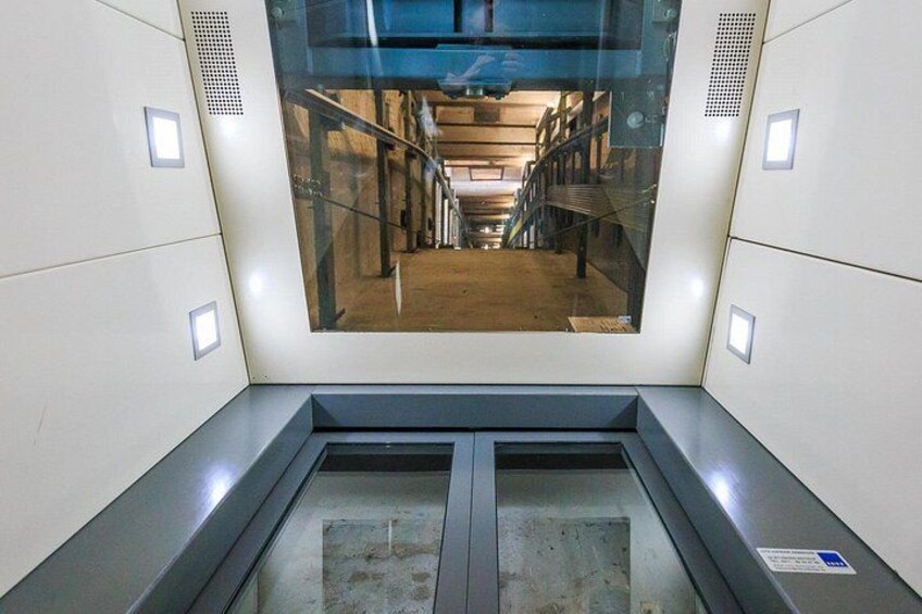 Een liftinterieur met een groot raam, metalen handlijsten en een verdieping aanduiding. 2