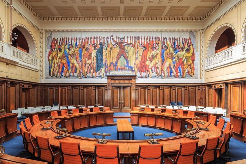 Un'ampia sala conferenze circolare con tavoli e sedie in legno, un tavolo centrale e un affresco raffigurante una scena storica sulla parete. 2