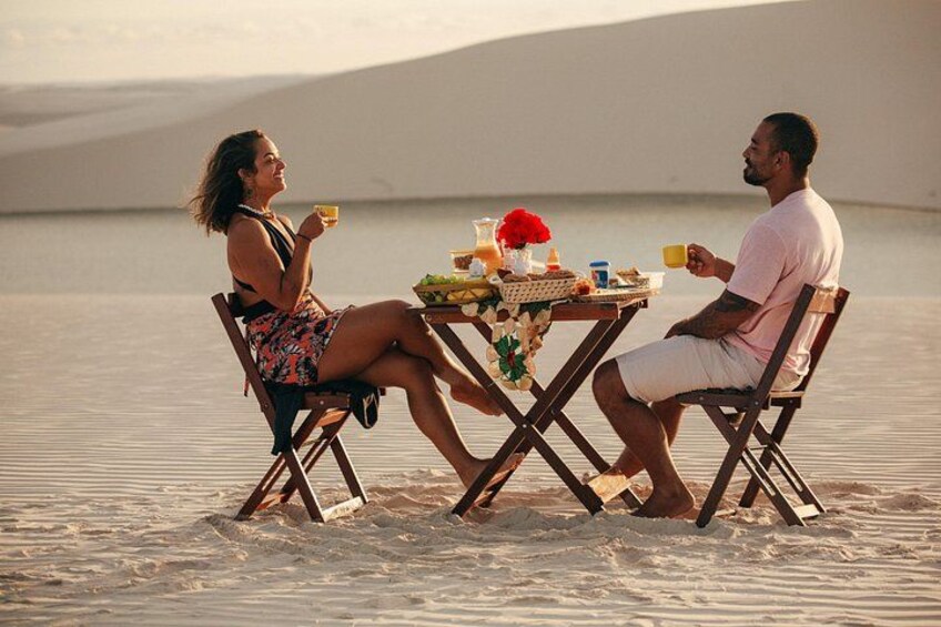 Two people sitting at a table on a sandy beach with a desert landscape in the background. 3