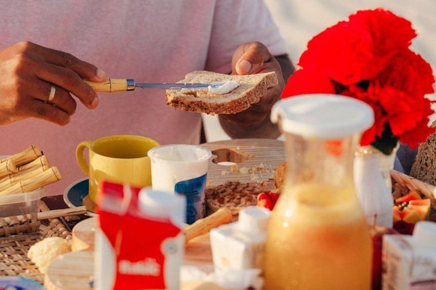 A person cutting toast with a knife, a yellow mug, a blue cup, a red container, and a pitcher of a yellowish liquid. 2