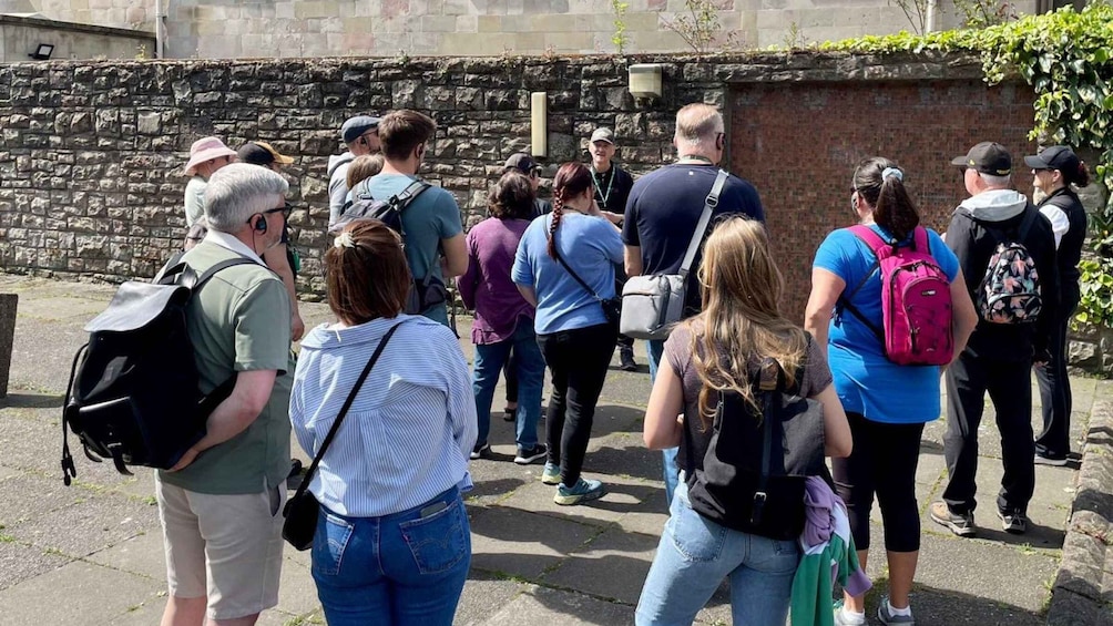 Un groupe de personnes se tient à l’extérieur, certaines ont des sacs à dos, et elles se trouvent près d’un mur de pierre. 4