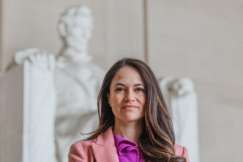 A person in a pink blazer with a purple shirt, standing in front of a white statue. 4