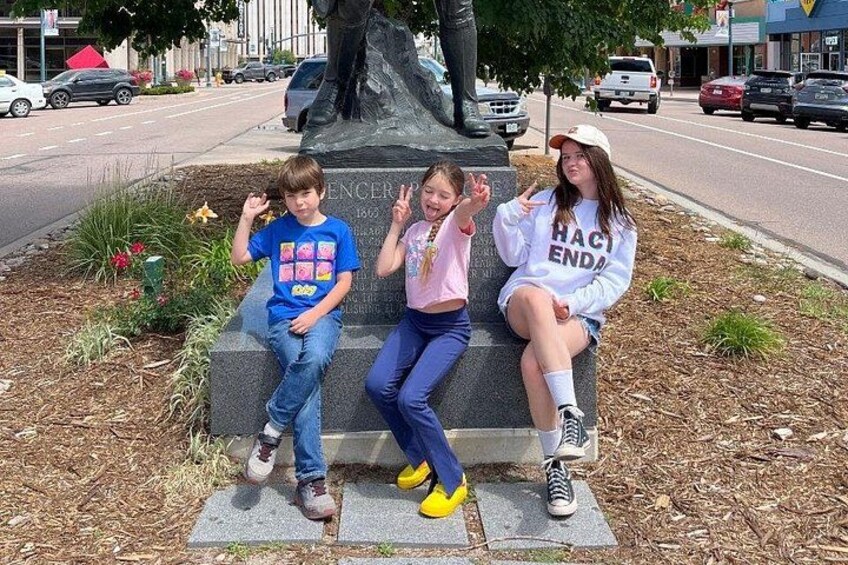 Three people sitting on a bench near a statue in a park with a road in the background. 2