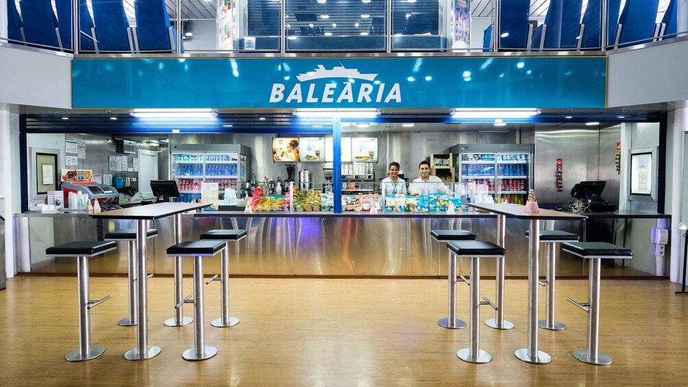 A BALEÁRIA food counter with a blue sign, two baristas, and a variety of snacks and drinks. 3