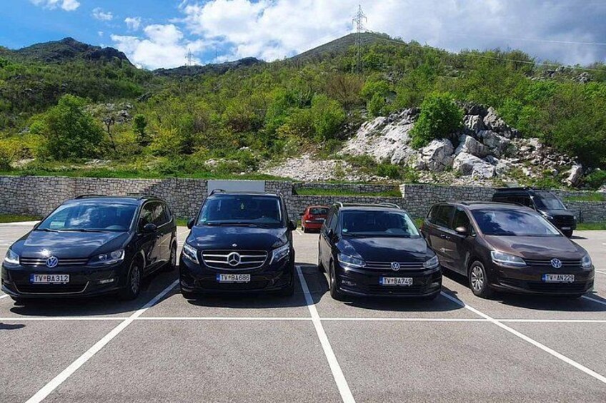 A parking lot with five cars, a stone wall, and a mountainous landscape in the background. 3