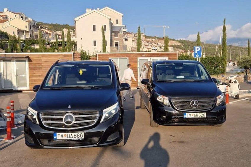 Twee zwarte Mercedes-Benz auto's staan op een parkeerplaats, met een gebouw en huizen op de achtergrond. 4