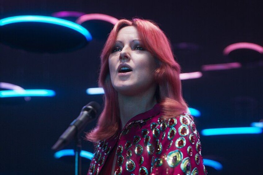 A person with red hair wearing a red and gold patterned outfit, standing in front of a microphone. 5
