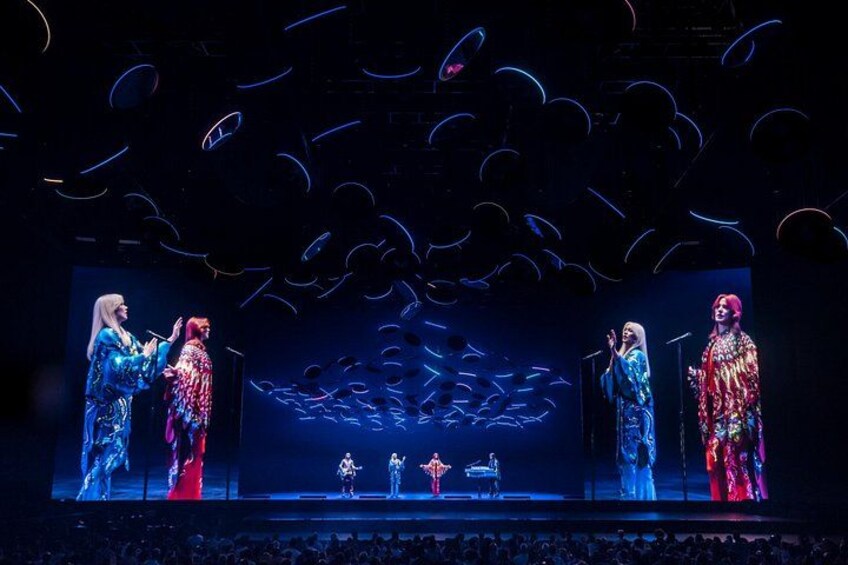 A stage with performers in colorful costumes, a large screen displaying abstract patterns, and a dark background. 4