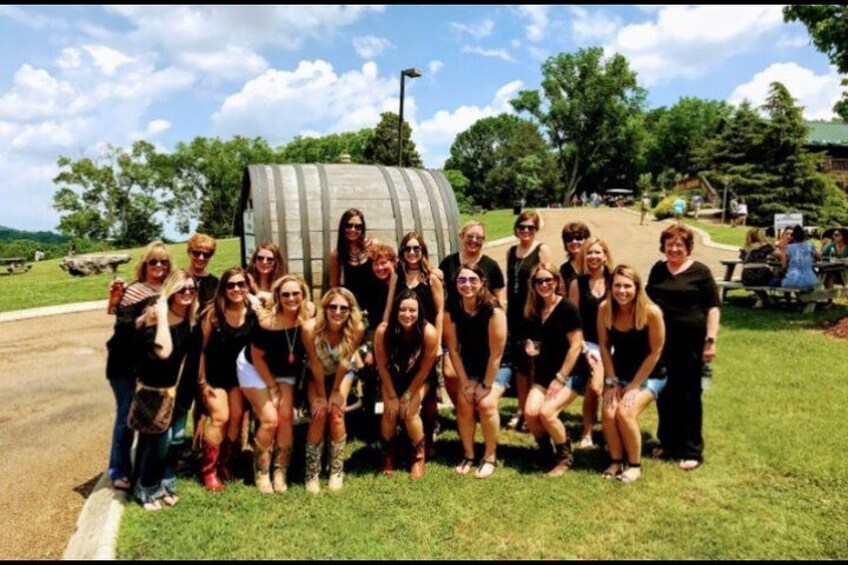 A group of people posing in front of a large barrel-like structure in a grassy area with trees and a clear sky in the background. 3