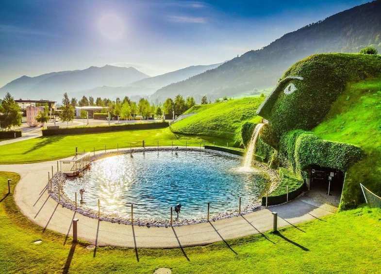 Ein kreisrunder Teich mit einem Wasserfall, umgeben von grünem Gras und einer Bergkulisse. 2
