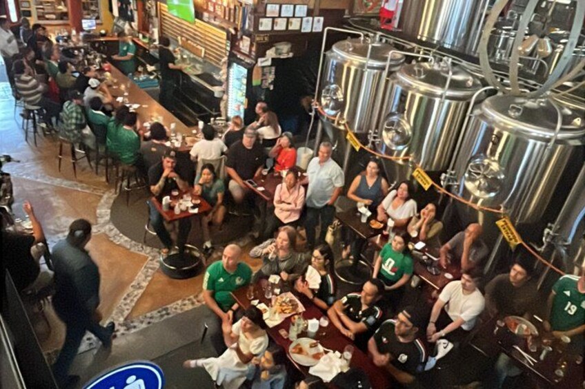 A crowded bar with people sitting at tables, surrounded by large stainless steel brewing tanks. 2