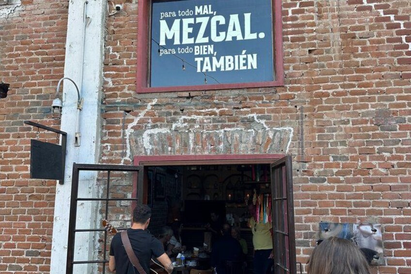 A brick building with a sign that reads 'MEZCAL. para todo MAL, para todo BIEN, TAMBIÉN' and a storefront with a metal gate. 3
