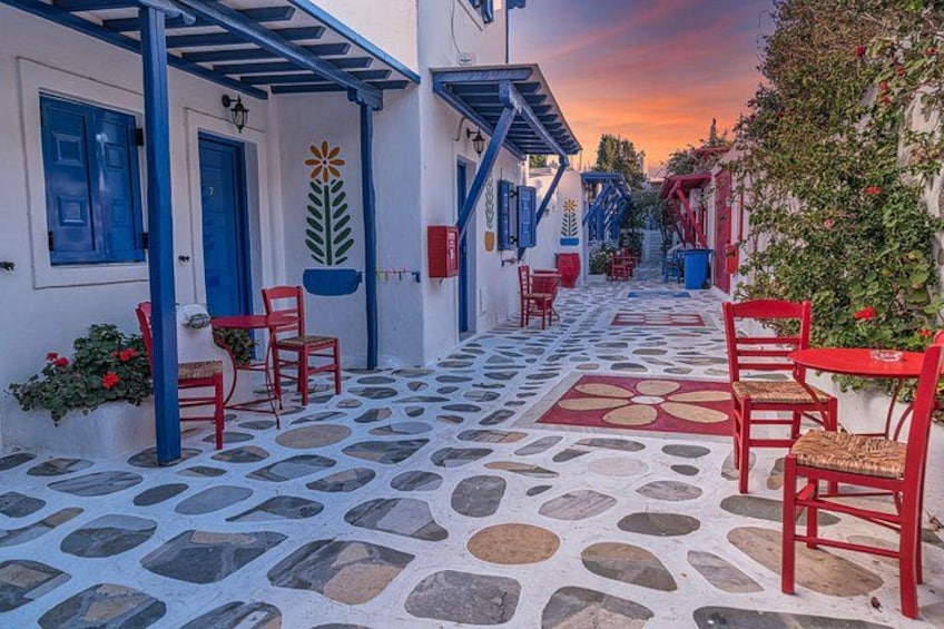 A narrow street with red chairs and tables, stone pavement, and blue doors. 4