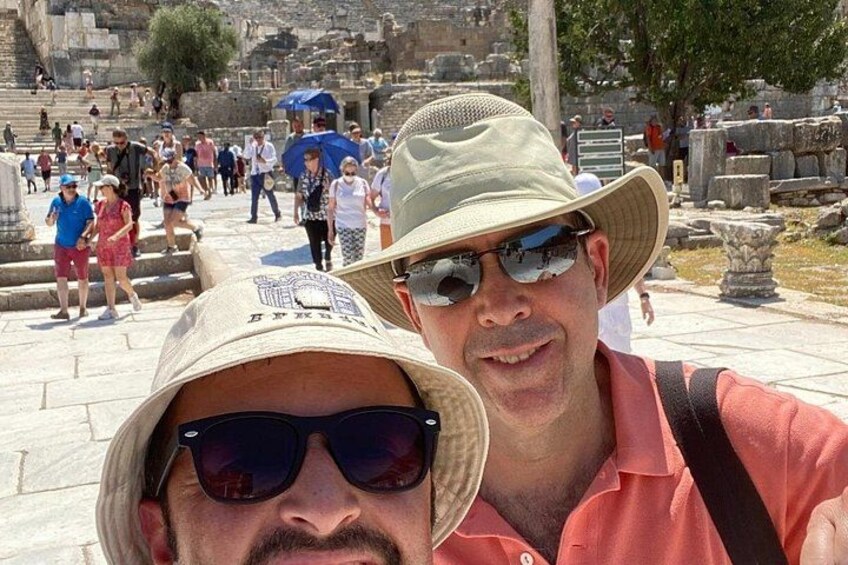 Zwei Personen machen ein Selfie auf einem historischen Gelände mit Ruinen und Besuchern im Hintergrund. 3