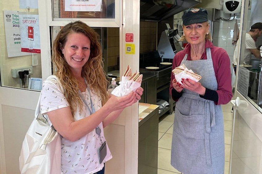 En person som håller i en smörgås och en väska, och som står i ett köksområde med rostfria köksredskap, med en annan person i bakgrunden. 1
