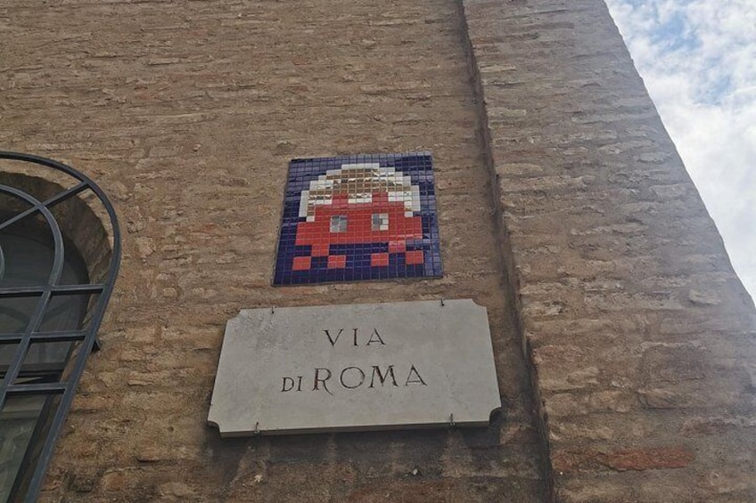 A mosaic of a face on a brick wall with a sign below that reads 'VIA DI ROMA'. 4