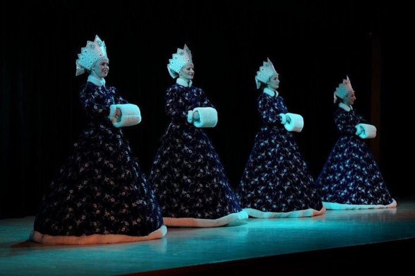 Four figures in dark blue dresses with floral patterns, standing in a row on a stage with a dark background. 3