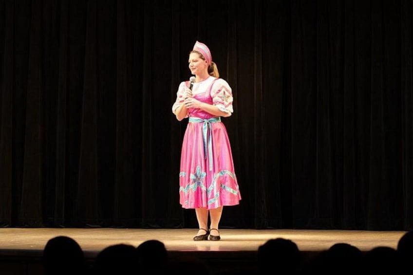 A person on stage wearing a pink dress with blue patterns, holding a microphone. 3