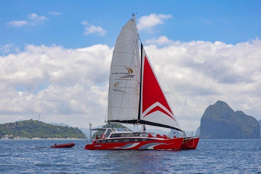 A red and white sailboat with a red sail, sailing on calm waters with a mountainous island in the background. 2
