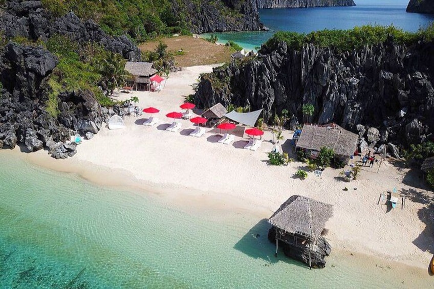 A beach with clear turquoise water, white sand, and red umbrellas. There are thatched-roof huts and rocky cliffs in the background. 4