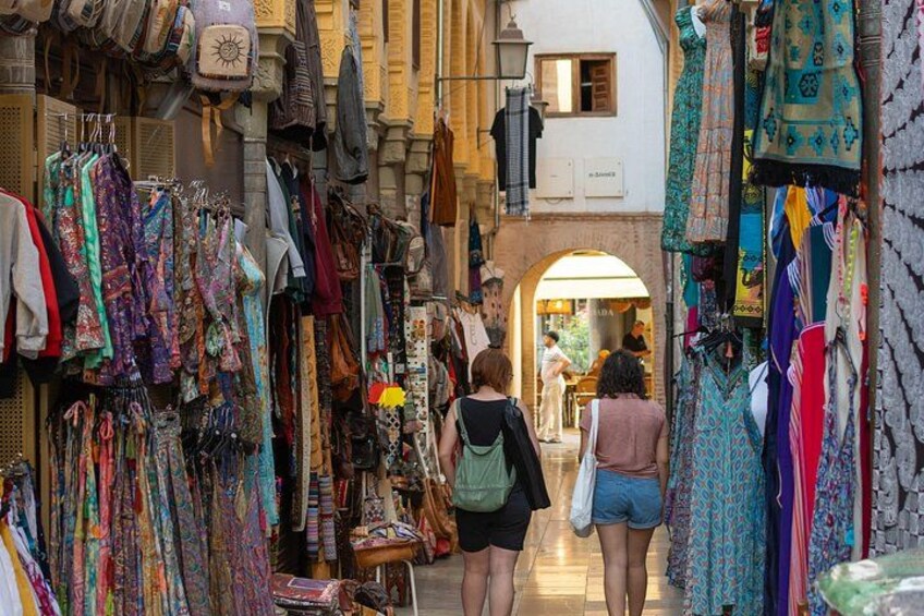 A narrow street with colorful textiles and clothing stalls. 1