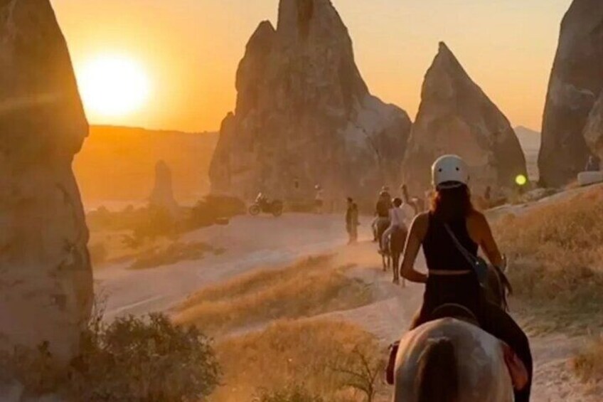 A group of people horseback riding on a trail with large rock formations and the sun setting in the background. 4